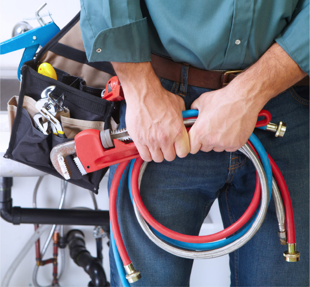 HVAC technician holding plumbing tools and flexible hoses in front of a tool bag and exposed piping.